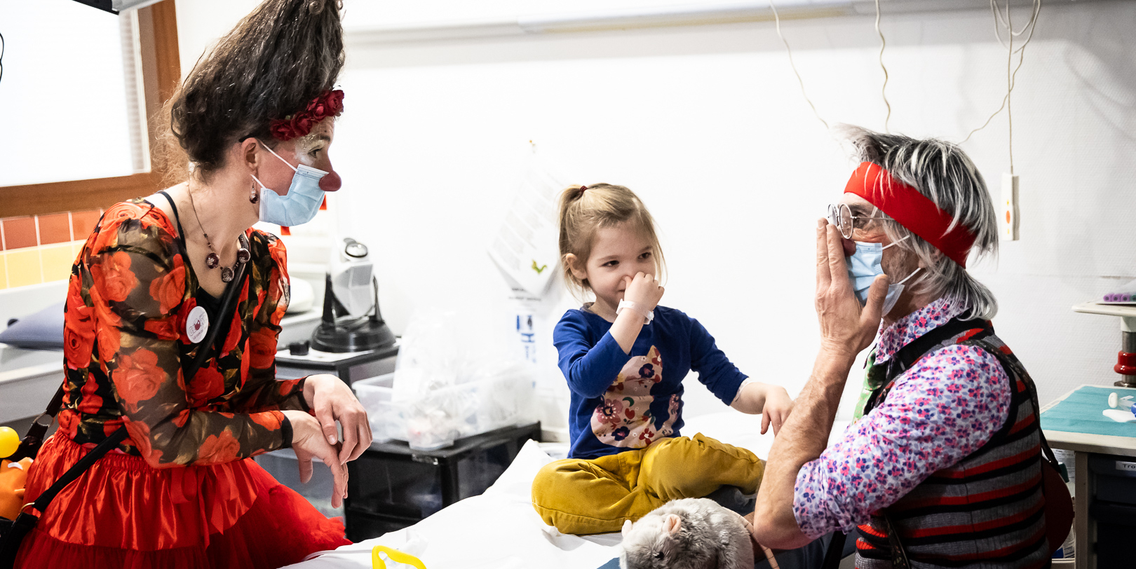 Deux clowns jouent avec une enfant dans une salle de soins.